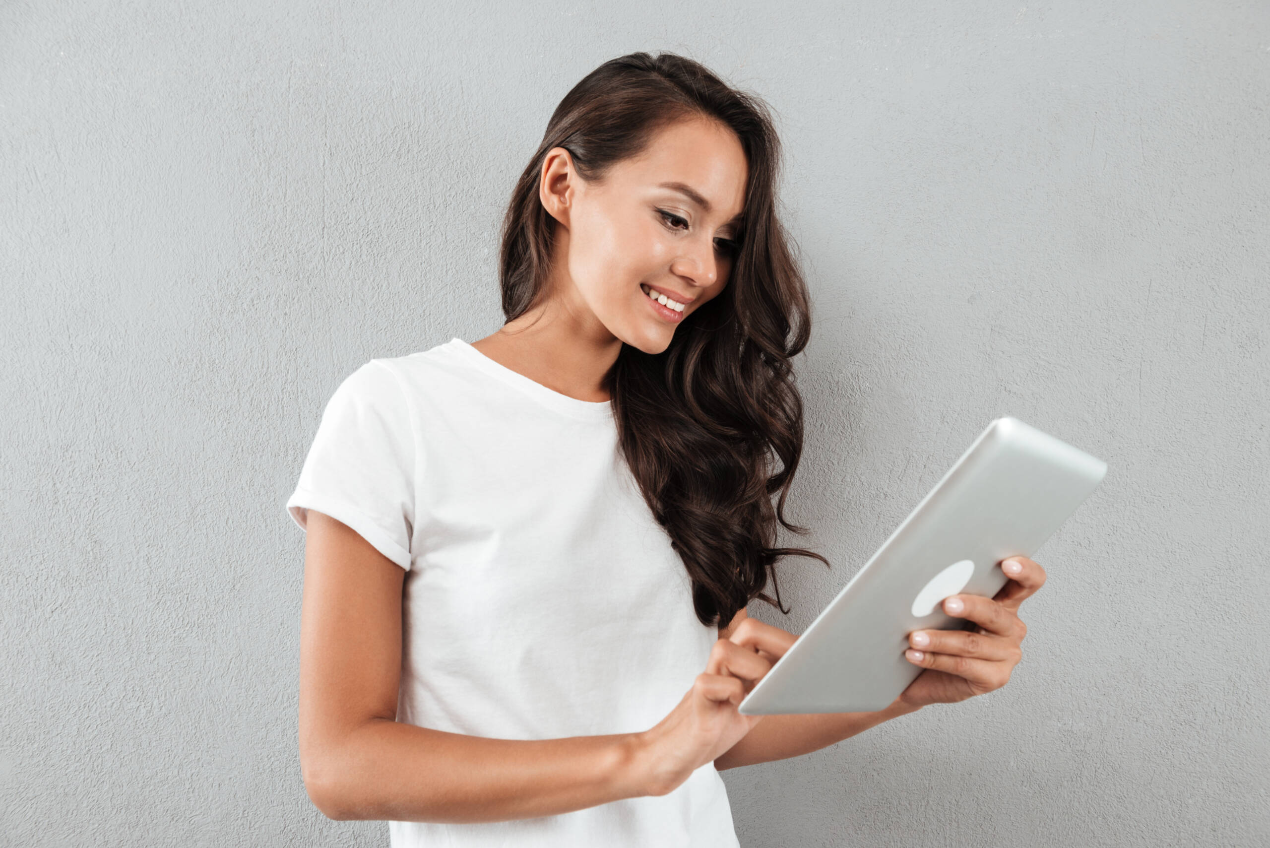 Smiling asian woman using tablet computer while standing isolated over gray background